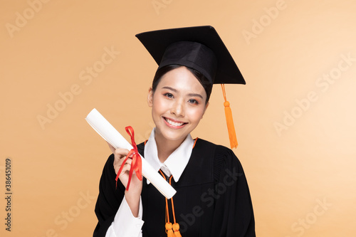 Happy Graduate asian woman in cap and gowm holding Certificated or diploma on Beige background,Graduation Concept photo