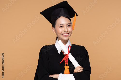 Happy Graduate asian woman in cap and gowm holding Certificated or diploma on Beige background,Graduation Concept photo