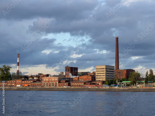 view of the old red brick factory buildings across the river against a cloudy sky illuminated by the rays of the setting sun