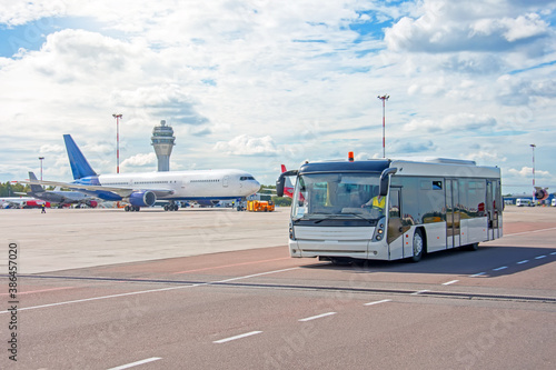 Bus for transporting passengers to boarding aircraft, against the background of a taxiing airliner on the apron of the airfield with a control tower.