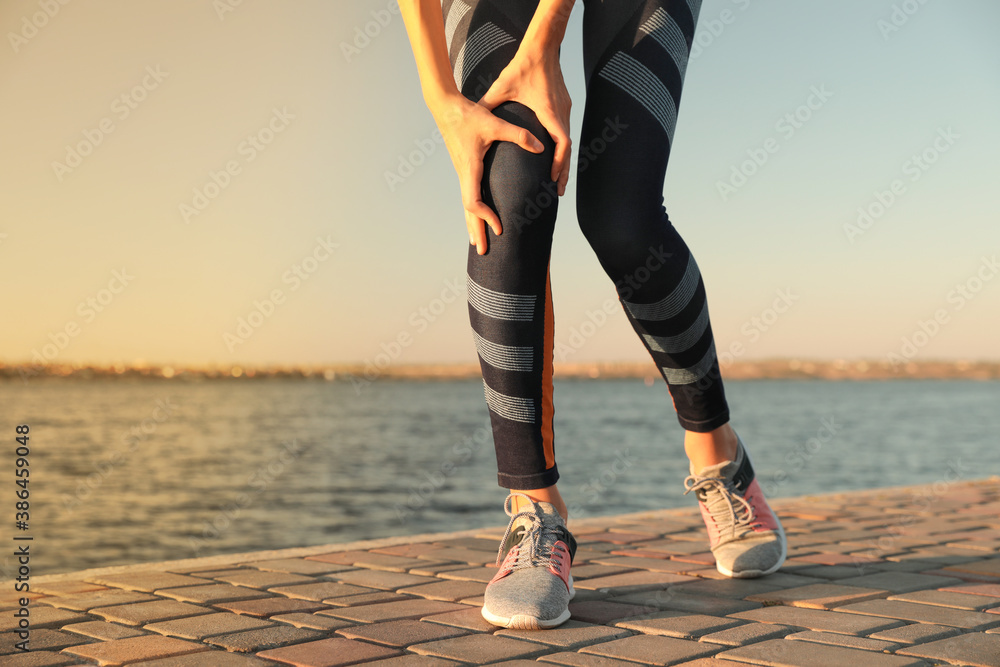 Young woman in sportswear having knee problems near river at sunset, closeup