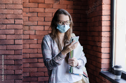 Girl student in a protective medical mask on her face looks at the phone. Communication by wireless technologies. Distance and self-isolation in a pandemic. photo