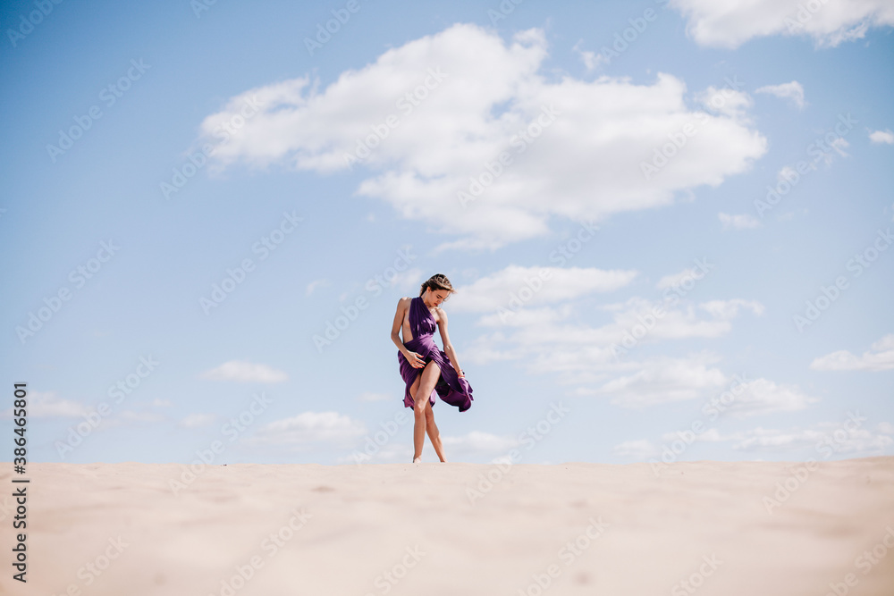 A young, slender girl in a beige dress with purple cloth in her hands posing in the desert in the wind