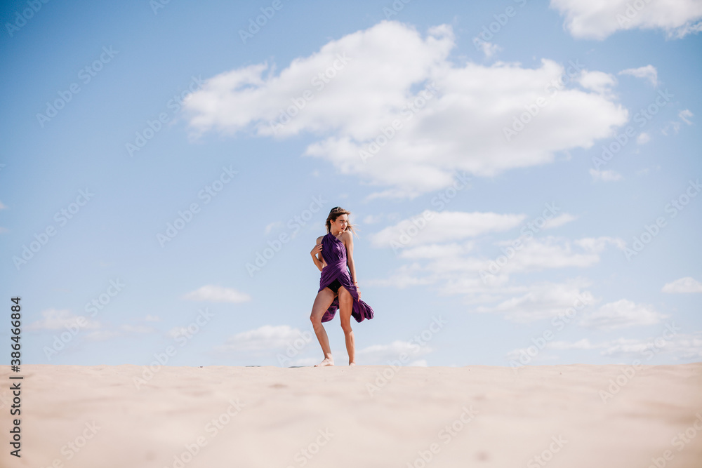 A young, slender girl in a beige dress with purple cloth in her hands posing in the desert in the wind