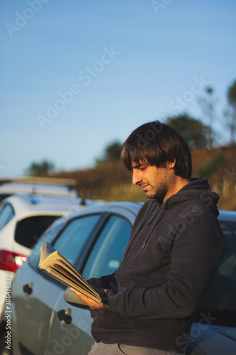 Young man reading a book leaning on a parked car in the sunlight with trees at the back and clear skies