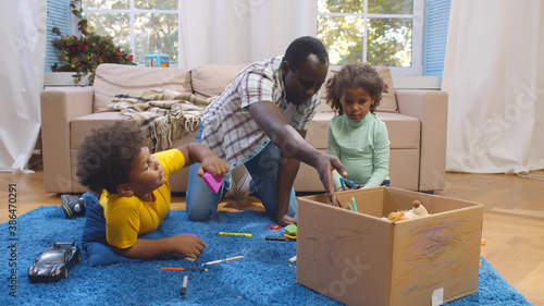 Quadro su tela African father and two preschool children sitting on carpet and putting toys in