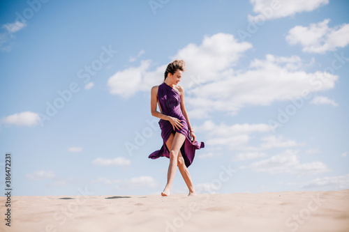 A young, slender girl in a beige dress with purple cloth in her hands posing in the desert in the wind
