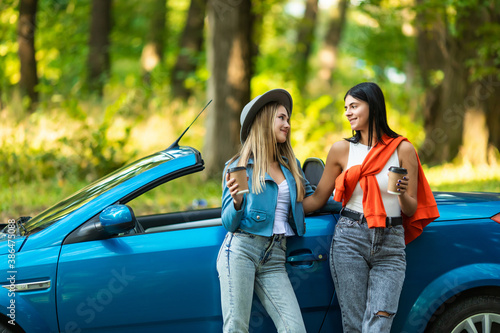 Two young women friends drinking take away coffee while standing near convertible car on the round