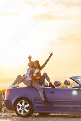 Two women sitting on the trunk of convertible car have fun time and enjoy the sunset