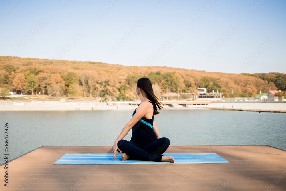sportswoman performs stretching on a blue Mat on the pier against the background of the lake