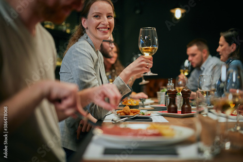 Charming smiling woman sitting in restaurant with friends on dinner and holding glass of white wine.
