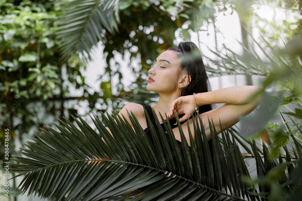 Beautiful mixed raced Asian woman model with dark long ponytail hair style, wearing black dress, posing in greenhouse with tropical plants, standing behind palm tree leaf. Tropical, summer concept. Beautiful mixed raced Asian woman model with dark long ponytail hair style, wearing black dress, posing in greenhouse with tropical plants, standing behind palm tree leaf. Tropical, summer concept.