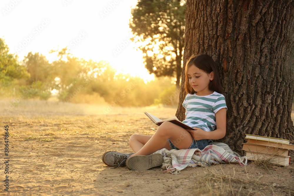 Cute little girl reading book near tree in park - obrazy, fototapety, plakaty 
