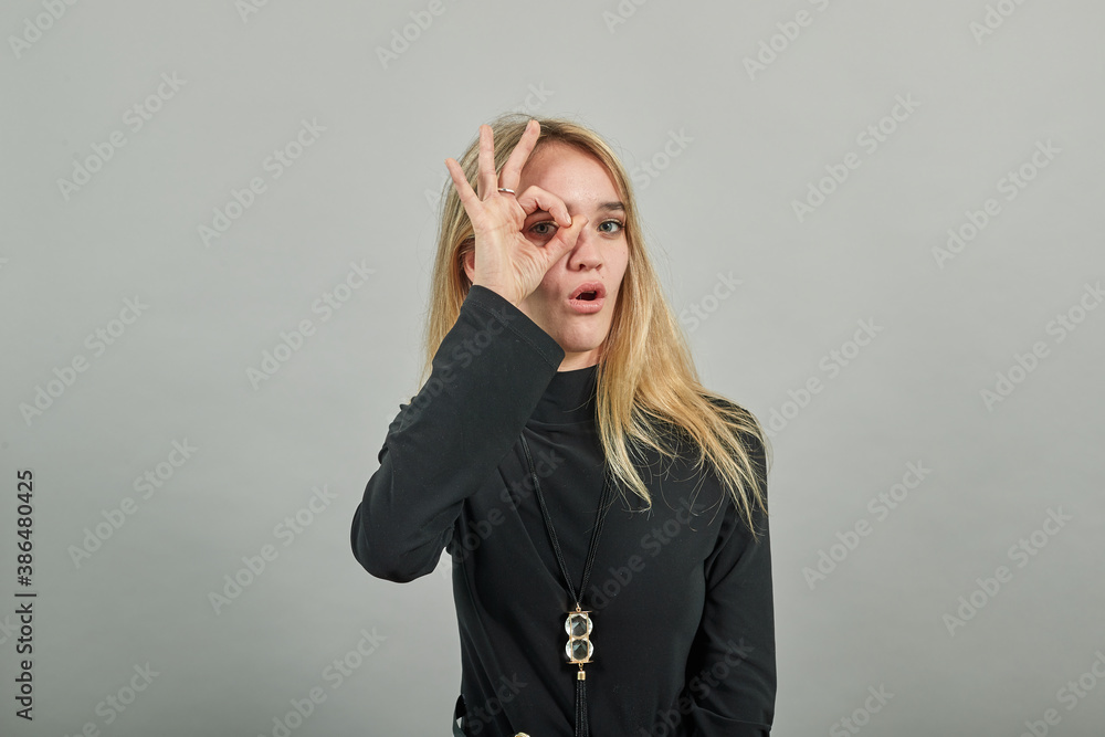 View of hand fingers showing ok sign, gesturing making okay, human emotions, facial expressions, feelings. Young attractive woman, dressed black sweater with green eyes, blonde hair, background