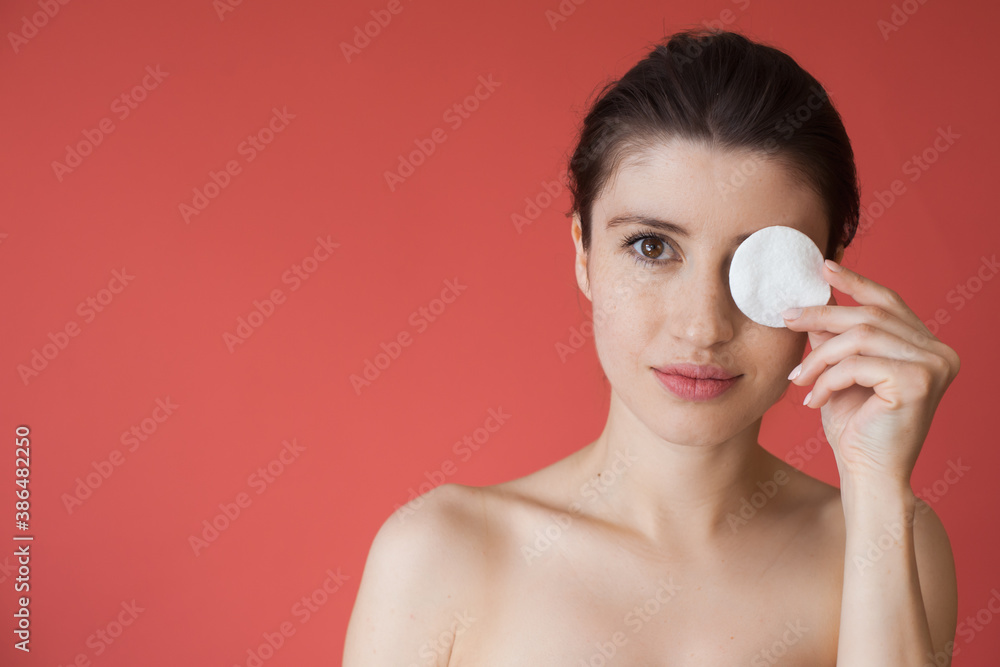 Freckled caucasian woman is covering her eye with a cotton disc smiling on a red wall with naked shoulders Freckled caucasian woman is covering her eye with a cotton disc smiling on a red wall with naked shoulders