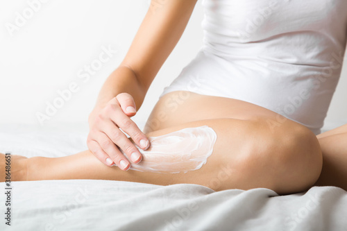Young adult woman sitting on bed and applying white moisturizing cream on leg. Daily care about clean and soft body skin. Closeup. Front view.