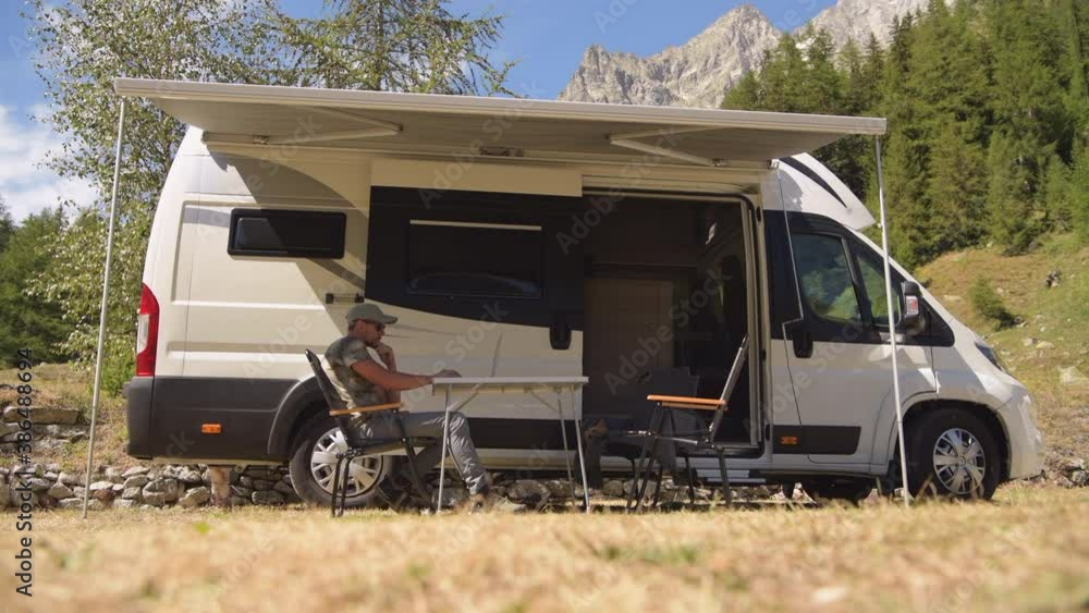 Caucasian Men Browsing Internet Outside of His RV Motorhome on a Camping. Scenic Camping Pitch. Caucasian Men Browsing Internet Outside of His RV Motorhome on a Camping. Scenic Camping Pitch.