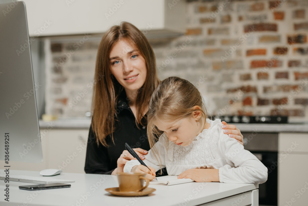 A mom is posing and hugging a pretty daughter while she is drawing in the notebook at the workplace. A gorgeous mother watches her blonde child which is doing homework at home. A mom is posing and hugging a pretty daughter while she is drawing in the notebook at the workplace. A gorgeous mother watches her blonde child which is doing homework at home.