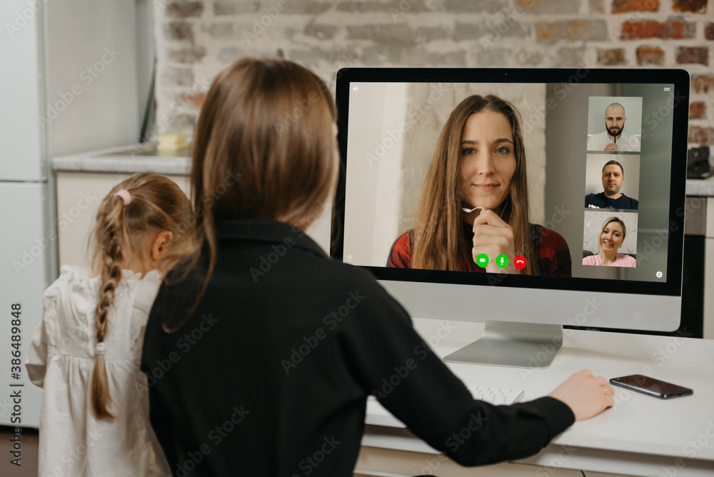 A young mother during a video conference with colleagues on the desktop computer while her daughter is drawing. A gorgeous mom is working remotely with her blonde child at home A young mother during a video conference with colleagues on the desktop computer while her daughter is drawing. A gorgeous mom is working remotely with her blonde child at home