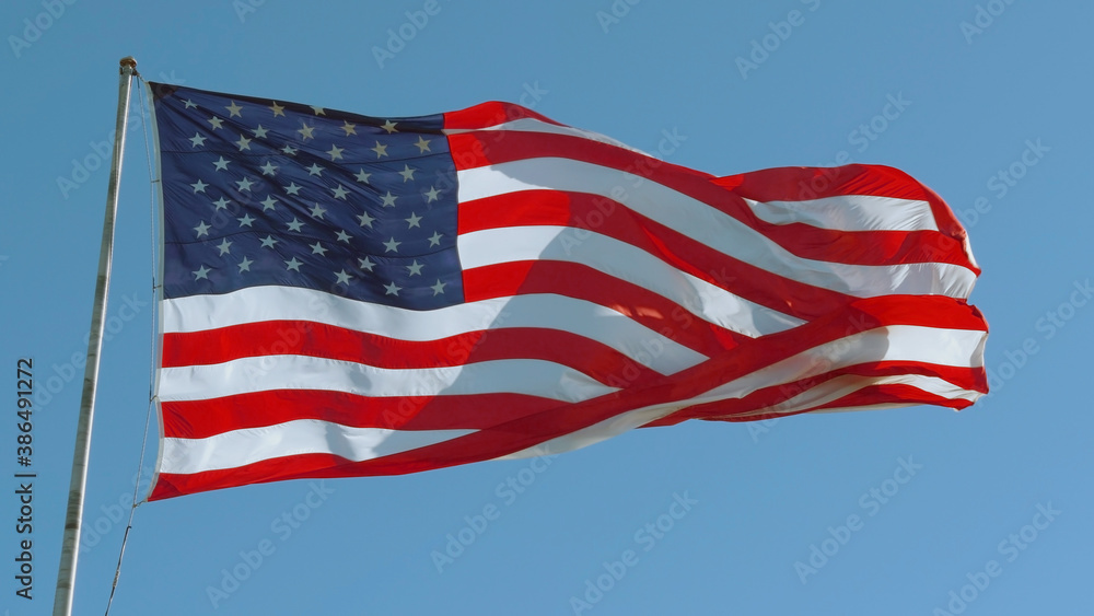 Fototapeta premium American USA flag on a flagpole waving in the wind against a clear blue sky on a sunny day.