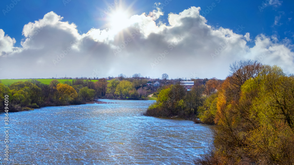 Spring landscape with a river and a picturesque cloud in the blue sky through which the sun looks