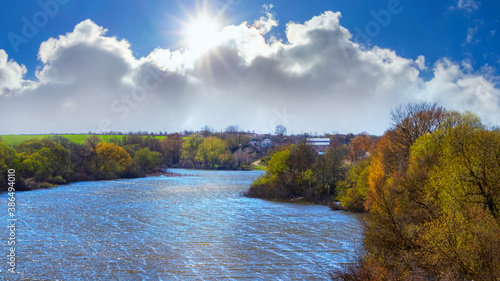 Spring landscape with a river and a picturesque cloud in the blue sky through which the sun looks
