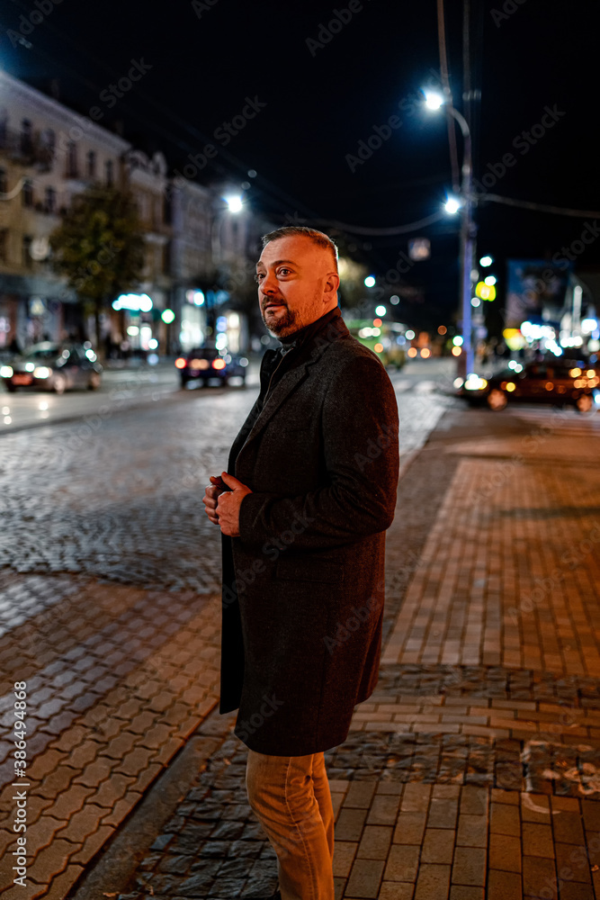 Mature fashionable adult man dressed in coat walking in urban setting. Stylish man walking on the street in the evening.