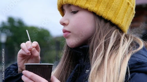 little girl in a jacket and a hat drinks hot coffee / hot chocolate / cocoa on the street on an autumn day