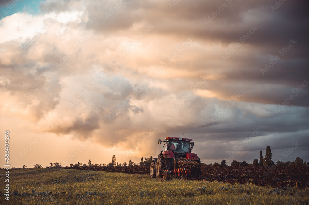 Tractor plowing the fields in the countryside by sunset. Agricultural tractor plowing the field. Red Tractor with plow. Sunset over the autumn field. Tractor plowing the fields in the countryside by sunset. Agricultural tractor plowing the field. Red Tractor with plow. Sunset over the autumn field.