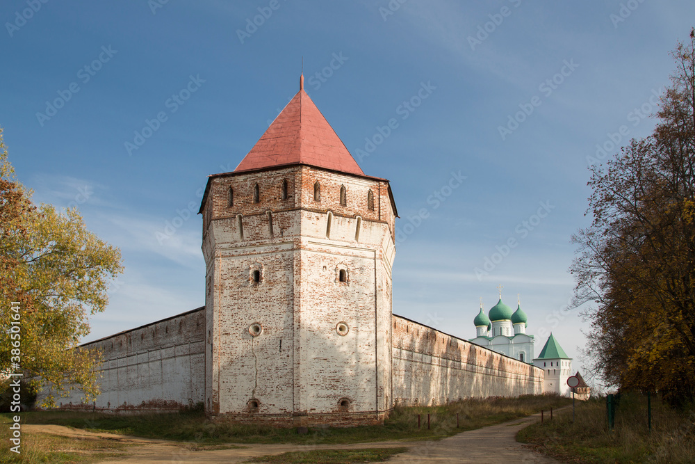 A whitewashed tower with visible red bricks and a wall around a Russian monastery. White church with green domes in the background. Yellow and red trees around. Blue sky. No people.