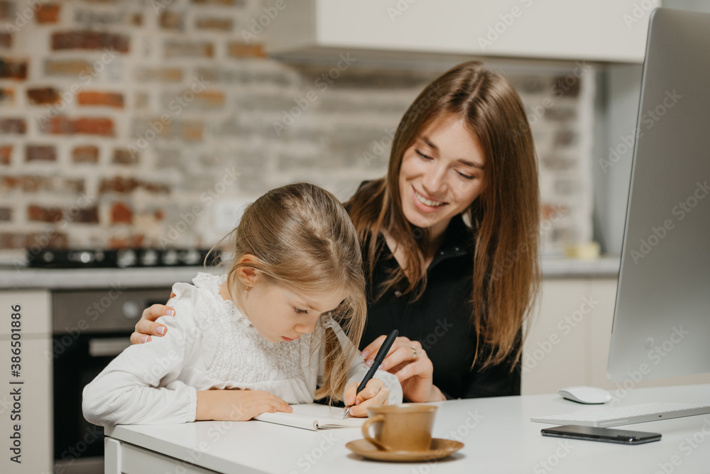 A young smiling mom is hugging a pretty daughter while she is writing in the notebook at the workplace. A gorgeous mother is looking at her blonde child which is doing homework at home. A young smiling mom is hugging a pretty daughter while she is writing in the notebook at the workplace. A gorgeous mother is looking at her blonde child which is doing homework at home.