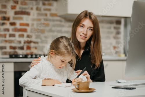 A mom is posing and hugging a pretty daughter while she is drawing in the notebook at the workplace. A gorgeous mother watches her blonde child which is doing homework at home.