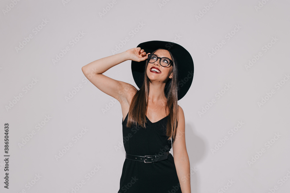Exited stylish happy girl wearing black dress and hat holding glasses and looking up with smile over isolated background Exited stylish happy girl wearing black dress and hat holding glasses and looking up with smile over isolated background