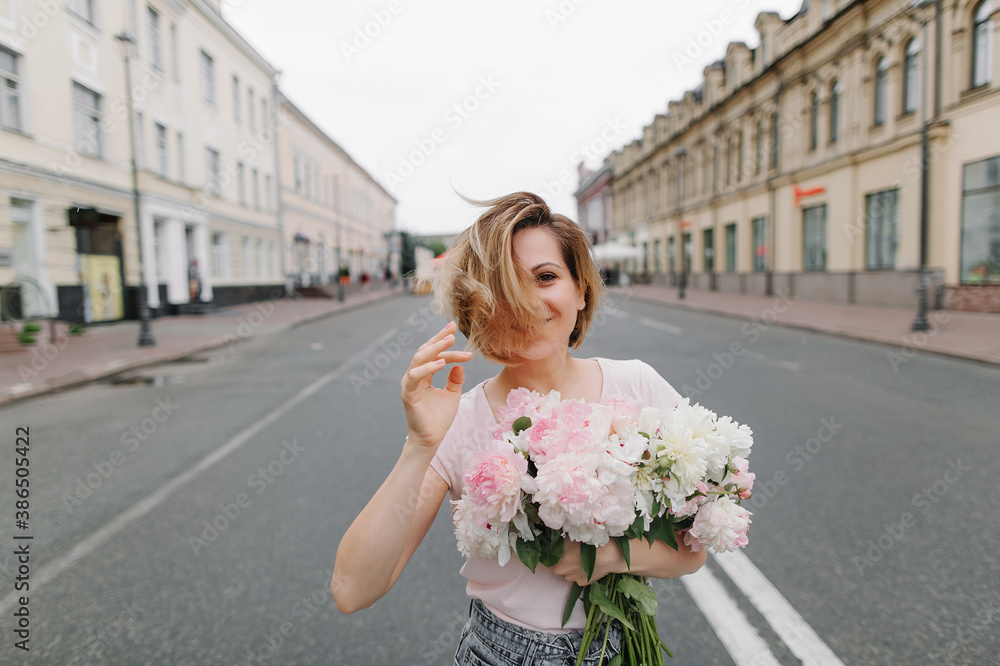 Fototapeta premium Happy girl with a bouquet of peonies walking through a deserted city in the early morning