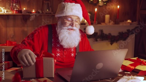 Old Santa Claus holding gift box video calling kid talking to child in virtual video online chat meeting on laptop sit at home table late saying greetings on xmas eve. Merry Christmas social distance. photo