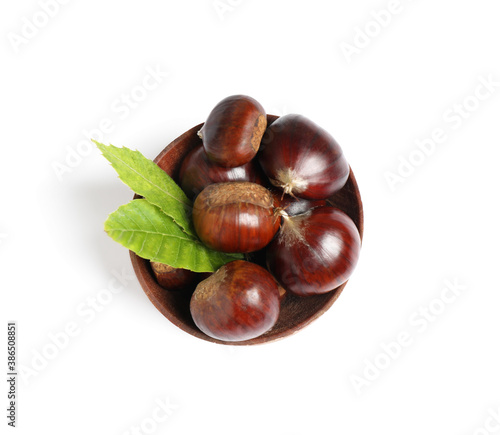 Fresh sweet edible chestnuts with green leaves in wooden bowl on white background, top view