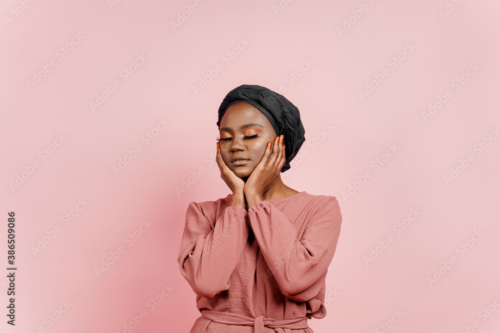 Muslim young woman weared in traditional dress and scarf touches her face and relax. Attractive and confident black model in studio isolated on pink background. Relaxation and meditation with yoga Muslim young woman weared in traditional dress and scarf touches her face and relax. Attractive and confident black model in studio isolated on pink background. Relaxation and meditation with yoga