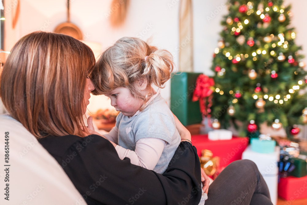 Mother and daughter cuddling on the sofa, Christmas time scene with tree and gifts on background - Woman and little girl on the sofa hugging and playing in lockdown because of coronavirus Mother and daughter cuddling on the sofa, Christmas time scene with tree and gifts on background - Woman and little girl on the sofa hugging and playing in lockdown because of coronavirus