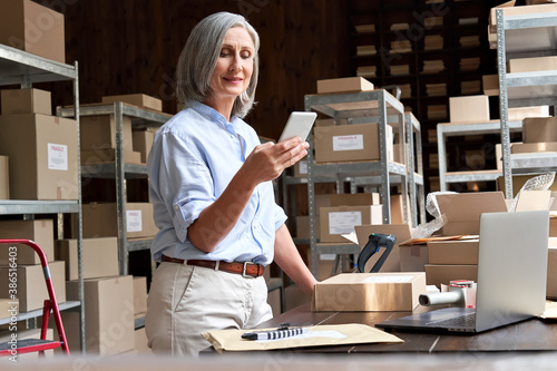 Female mature senior small business owner using mobile app checking parcel box. Older entrepreneur seller holding cell phone tech device preparing retail package postal shipping order in warehouse.