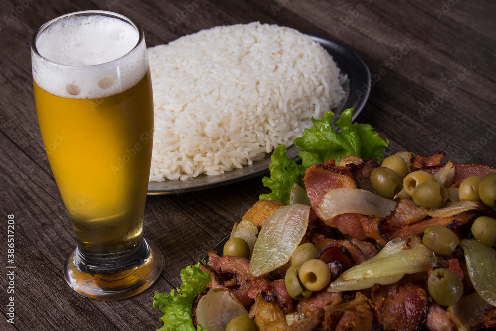 A refreshing beer accompanied by a portion of rustic fried chicken with rustic onions, bacon and a portion of white rice. Brazillian tipical food. Horizontal close-up gastronomic photography.