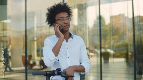 Young african agent with electric scooter talking on mobile phone in urban environment
