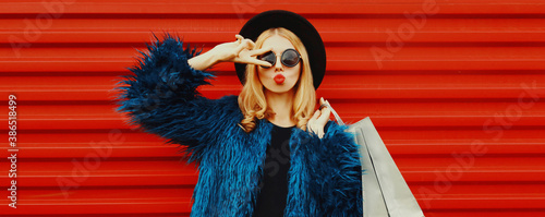 Portrait of attractive woman model with shopping bags blowing red lips sending sweet air kiss wearing blue faux fur coat, round hat over red wall background © rohappy