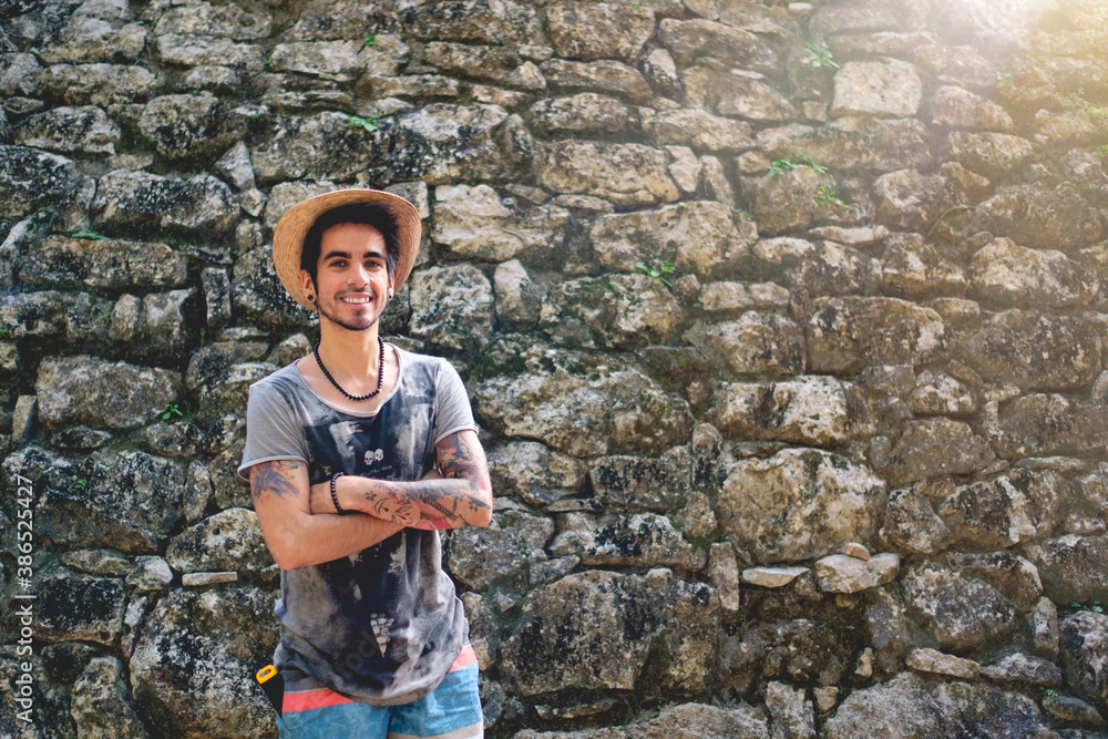 Young, tattooed and smiling traveler with hat in front of an old stone pyramid ruins at the Mayan archeological site at Coba, Mexico Young, tattooed and smiling traveler with hat in front of an old stone pyramid ruins at the Mayan archeological site at Coba, Mexico