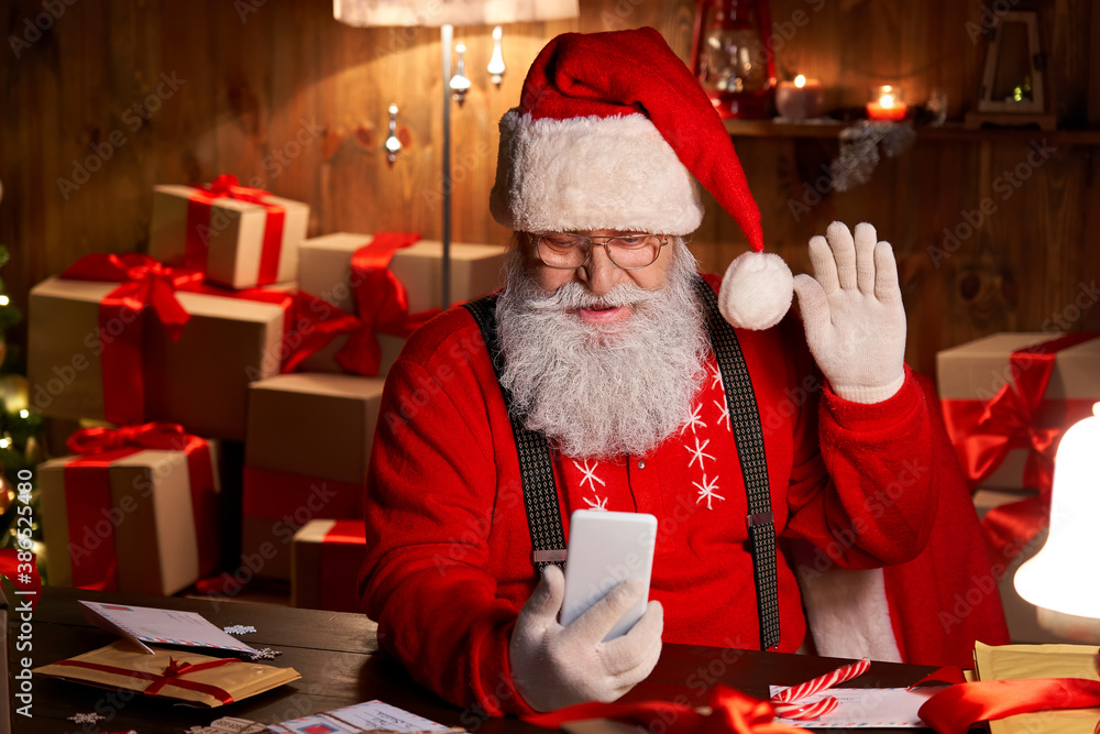 Φωτογραφία Happy Santa Claus holding smartphone video calling talking conferencing in virtual video online chat meeting sit at home table late with presents on xmas eve
