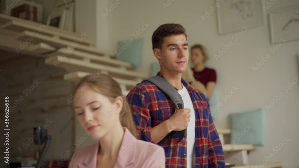 Tracking slow motion shot of young man with backpack entering office and discussing project on laptop with male colleague sitting at desk Tracking slow motion shot of young man with backpack entering office and discussing project on laptop with male colleague sitting at desk