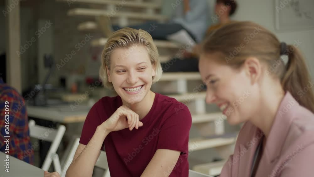 Tilt up medium shot of two young women in casual clothes discussing project on laptop computer cheerfully and laughing sitting at desk in office Tilt up medium shot of two young women in casual clothes discussing project on laptop computer cheerfully and laughing sitting at desk in office