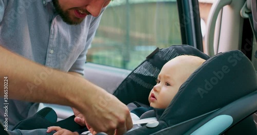 Camera inside the car. Closeup baby boy sits in the baby car seat inside of car. Young bearded father checks baby's fastened seat belts, and talks to him. Slow motion photo