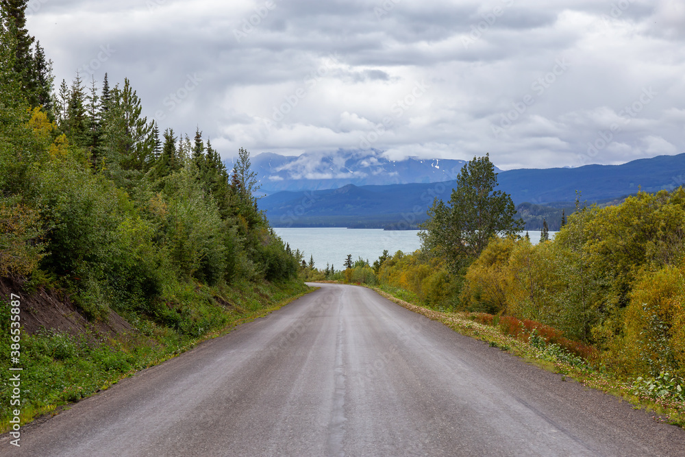 Fototapeta premium View of Scenic Road alongside Lake, surrounded by Trees and Mountains on a Cloudy Fall Day in Canadian Nature. Taken in Northern British Columbia, Canada.