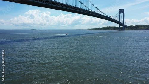 a low angle drone view of Lower New York Bay. The drone dolly in & pan right below The Verazzano Bridge with a single boat racing by. The sky is blue on a bright & beautiful day. photo