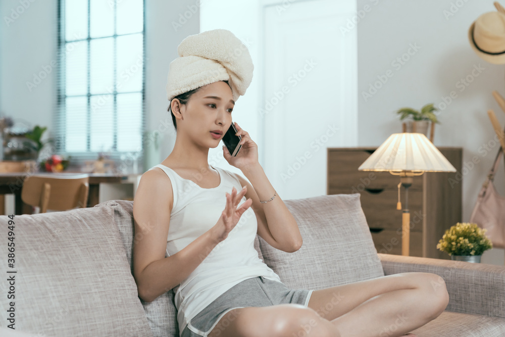 chinese female in casual wear is talking on phone in living room after taking bath. asian businesswoman hair wrapped in bath towel holding smartphone is explaining work with hand gesture.